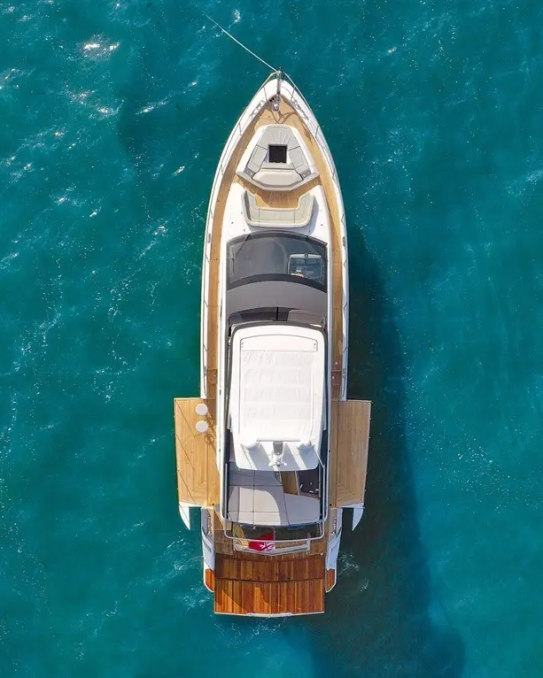 Aerial view of a luxury yacht with a wooden deck, moored in clear blue water, featuring a white sunbathing area and a flag.